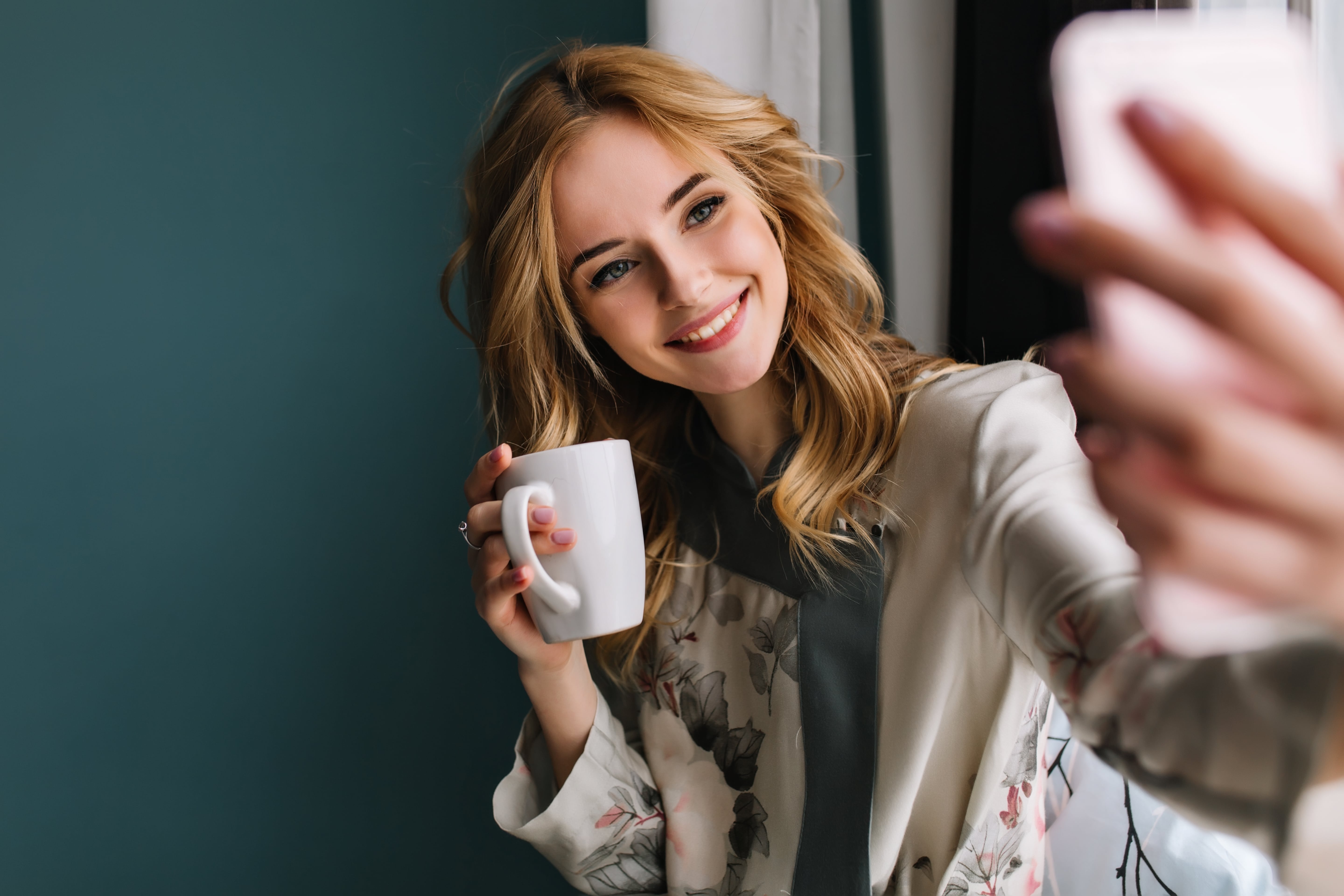 pretty-young-woman-with-wavy-blonde-hair-taking-selfie-sitting-window-with-cup-morning-coffee-tea-she-wearing-silk-pajama-turquoise-wall-min